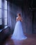 Lady in long blue vintage dress with train and open back stands with back to camera. room with large windows with