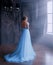 Lady in long blue vintage dress with train and open back stands with back to camera. room with large windows with