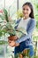 Lady long hair white t shirt light blue clothes holding Aglaonema tree stand smile look camera in glass room surround house plants