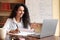 Lady sitting at table, using computer and writing in notebook