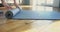 Lady yoga trainer unrolls grey rubber mat on wooden parquet