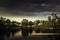 Lake with a small wooden dock on it surrounded by trees under a dark cloudy sky in the evening