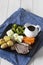 Lamb roast Sunday lunch dinner with vegetables on a aquare black plate, with gravy jug, on a blue tea towel.