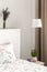 Lamp above cabinet with plant next to bed with sheets in bedroom interior with lavender flowers. Real photo