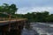 Landscape composed of a cloudy sky, riverside vegetation and a wooden bridge.