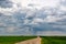 Landscape with dark sky with rain clouds before storm in green field with gravel road. thunderstorm front