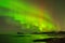 A landscape photograph with water and mountains, with a beautiful green Northern Light display in a dark star studded sky reflecti