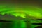 A landscape photograph with water and mountains, with a beautiful green Northern Light display in a dark star studded sky reflecti