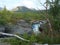 Landscape with rapids of wild blue glaciar river Kamajakka, rock, birch tree, green mountains and pink willowherb flowers at