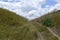 The landscape is a rustic view of a country road in the center between the hills and the blue cloudy sky