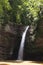 Landscape with summer hight waterfall in lush green forest with sunbeams, splashes and layered rocks overgrow green moss.