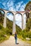 Landwasser Viaduct in summer, Filisur, Switzerland. Young woman looks at famous landmark of Swiss Alps. Red train runs on high