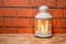 A lantern with a glowing candle in on the wooden table, brick wall background