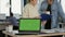 Laptop computer with green screen on desk in front of startup colleagues talking about sales charts papers