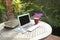 Laptop, face mask, books and red cup on top of table next to garden