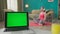 Laptop with a green screen are on the table in the living room. In the background, a woman in headphones sits in a lotus