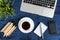 Laptop keyboard, white cup of tea on saucer, notepad, pen and green plant in the corner on dark blue crumpled jeans background.