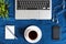 Laptop keyboard, white cup of tea on saucer, notepad, pen and green plant in the corner on dark blue crumpled jeans background.