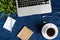 Laptop keyboard, white cup of tea on saucer, notepad, pen and green plant in the corner on dark blue crumpled jeans background.