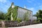 Large abandoned old stone family house partially overgrown on one side with crawler plants next to paved road surrounded with