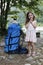 A large backpack for an adult stands next to a little girl against the backdrop of trees and a river, on a sunny day.