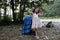 A large backpack for an adult stands next to a little girl against the backdrop of trees and a river, on a sunny day.