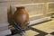 A large ceramic wine jug with copper vine decor sits on the floor of a cafe.