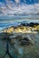 Large colourful rocks in calm shallow sea water soft waves blue sky with clouds, surfers in distance