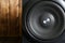 Large cubic subwoofer in a black wooden case with a metal grille stands against a background of natural pine boards. Audiophile