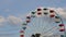 Large ferris wheel with open cabins quickly turns against a blue sky