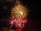 Large golden flash of fireworks and many small flashes in red with lilac sparks against a dark sky