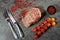 Large juicy ribeye steak with red pepper sprinkles in black saucer, rosemary sprigs and cherry tomatoes next to kitchen utensils