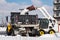 a large metal bucket of a tracked excavator loads snow from a city road onto a truck body