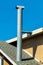 Large metal chimney vent with roof tiles and white accent color on tile edge with brown stucco exterior and blue sky