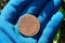 A large old copper coin lies on the palm of a hand in a blue glove