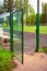 A large open metal door in a mesh fence behind which you can see a green football field and benches with a white canopy for