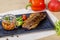 Large piece of grilled fish sits on a square black plate. Next to a jar of special sauce, a slice of lime, spinach, tomatoes.