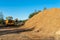 A large pile of wood chips lying on a square, with a blue sky in the background, visible bulldozer