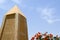 A large pointed obelisk made of yellow stone in Egypt against a blue sky and red flowers
