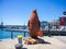 Large rusty penguin statue standing next to the bridge crossing at the Waterfront. Harbor and blue sky in background.