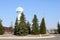 Large, tall, stone, concrete watch with arrows with a blue dial next to the big green Christmas trees on the background of an oil