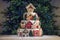 Large tiered Christmas cake decorated with gingerbread cookies and a house on top. Tree and garlands in the background.