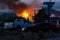 Large warehouse fire in the distance beyond spectators sitting in their cars shallow depth of field and focus