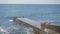 Large waves break on a concrete breakwater. A plump woman in a swimsuit lies on the breakwater getting a sun tan.