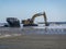 Large yellow excavator digs up sand at the edge of the ocean with a near by