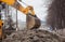 A large yellow excavator stands in the middle of the street near the dug hole