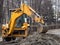 A large yellow excavator stands in the middle of the street near the dug hole