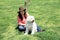 Latin adult woman with hat and white Labradoodle dog sitting in field, green grass outdoors