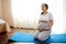 Latin American beautiful gravid woman holding her belly, doing breathing exercises on yoga mat at cozy homely atmosphere