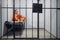 A Latin young criminal in a solitary cell reads a book sitting on a bunk in an orange prison uniform. Photo through the